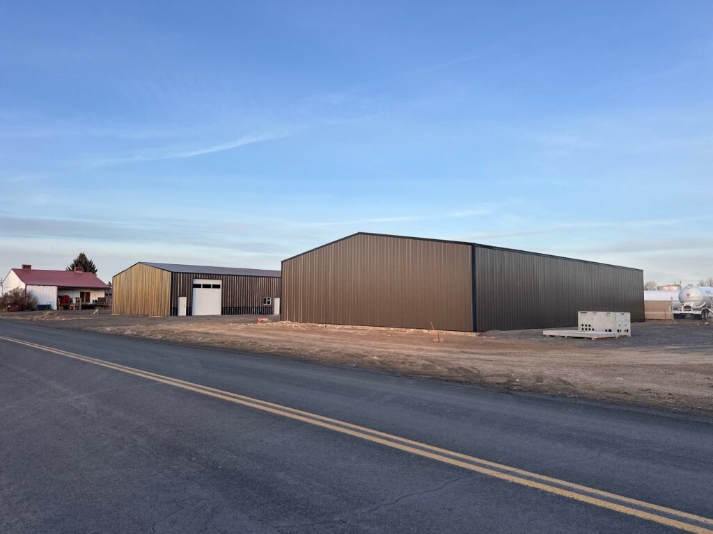Two pole buildings with metal siding and roof trusses, and and white garage doors, set near a red-roofed farm home.