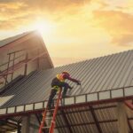 A worker repairs the metal roof of a commercial building while standing on a ladder at an active job site.