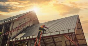 A worker repairs the metal roof of a commercial building while standing on a ladder at an active job site.