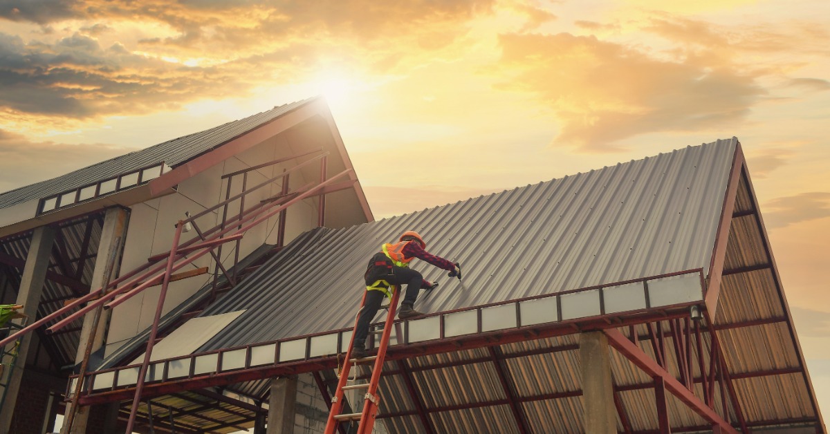 A worker repairs the metal roof of a commercial building while standing on a ladder at an active job site.