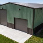 A green metal pole building with two overhead doors stands on a rural property surrounded by grass and trees.