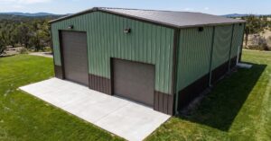 A green metal pole building with two overhead doors stands on a rural property surrounded by grass and trees.