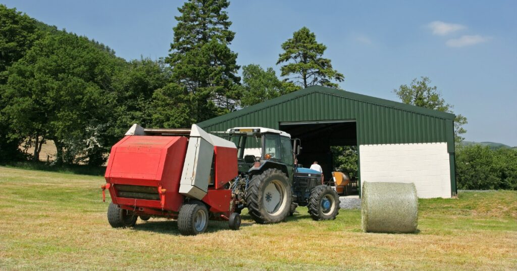 A tractor and hay baler sit beside the large open entrance of a green agricultural building on a rural property.
